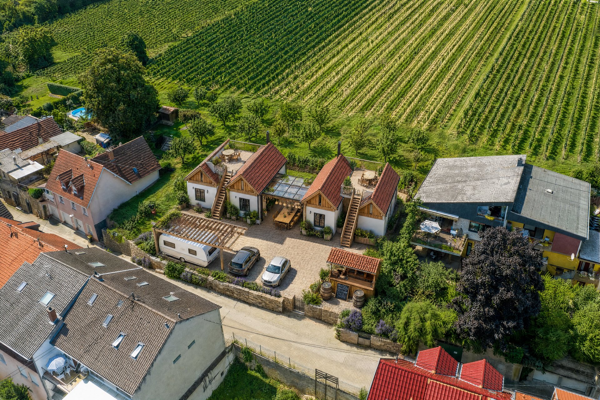 Variante C — Tiny-House-Compound von der Straßenseite mit Pavillon, Wohnwagen und Schank-Theke